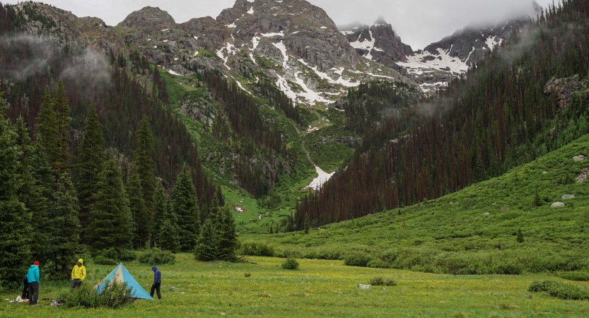 A tent sits in a green valley surrounded by mountains. Three people are standing near the tent.